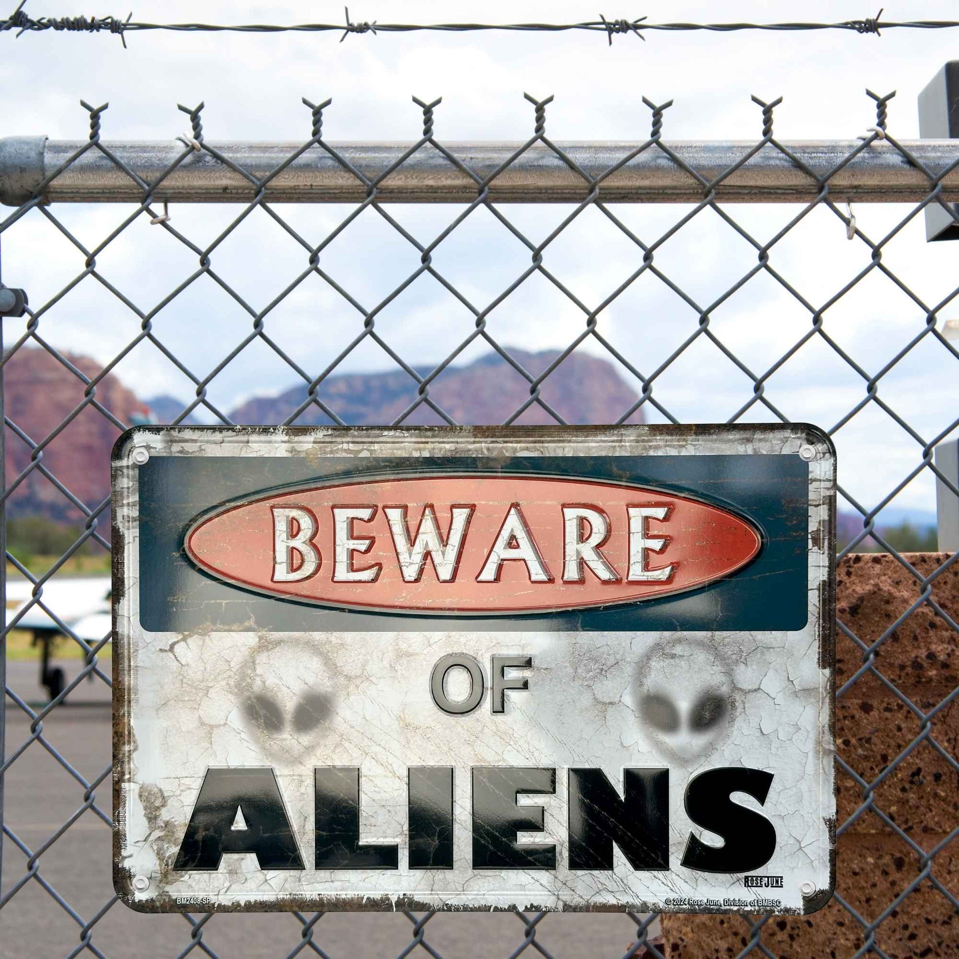 Vintage-style 'Beware of Aliens' sign behind a chain-link fence with desert landscape in the background.
