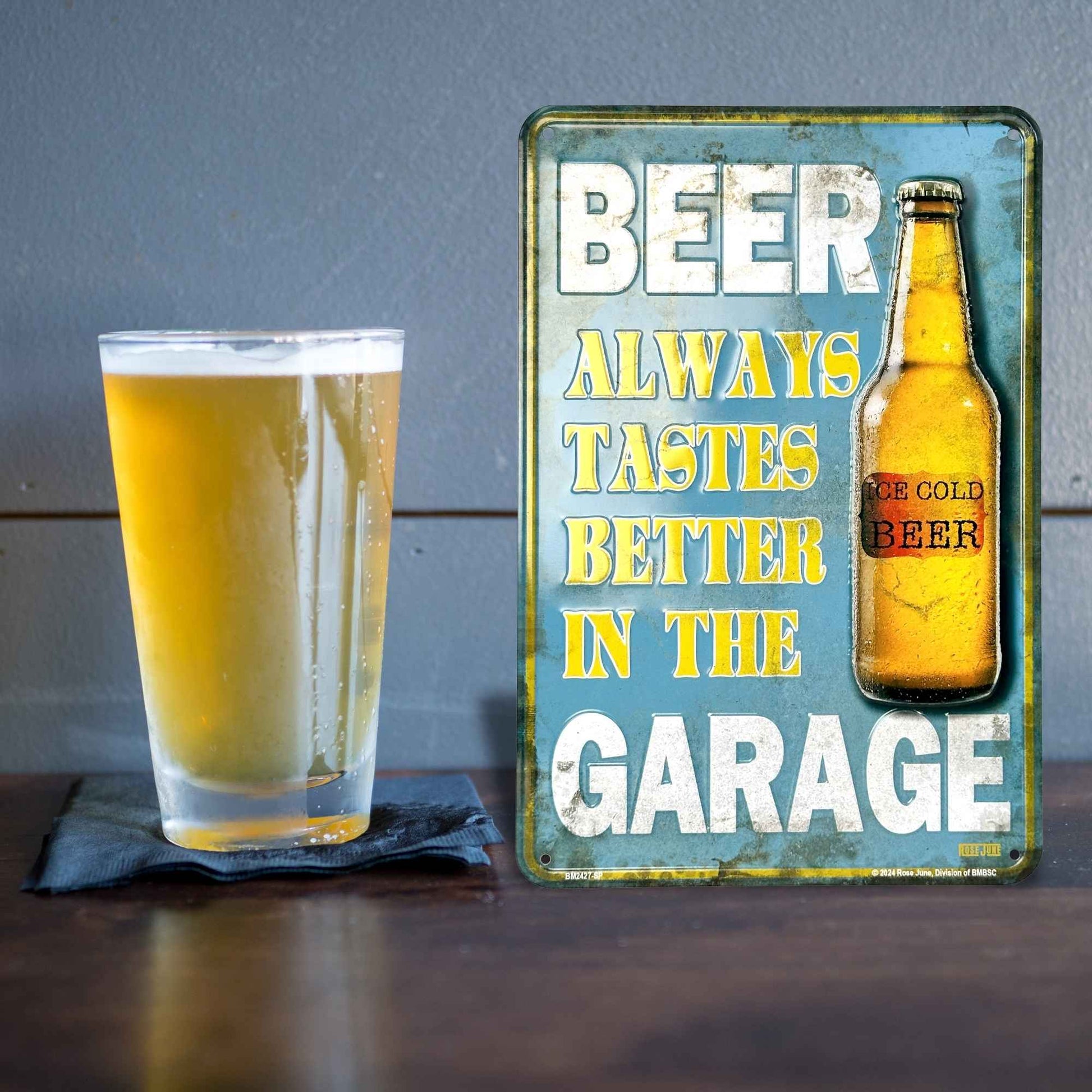 Glass of beer next to a metal sign with text and a beer bottle illustration on a wooden surface.