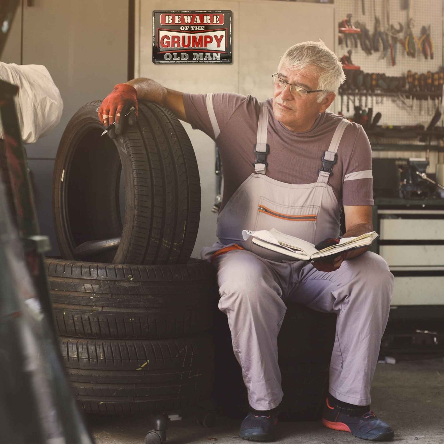 Man in a garage holding a tire and a book, with a 'Beware of the Grumpy Old Man' sign above.