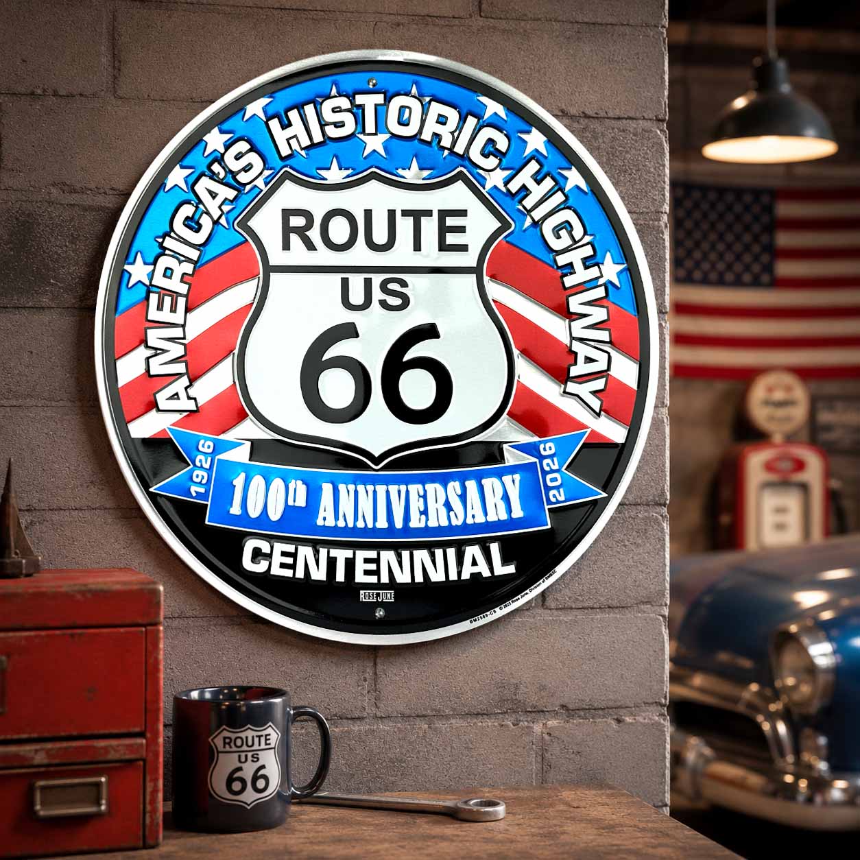 Decorative sign for Route 66 100th Anniversary Centennial on a brick wall with a vintage car and American flag in the background.